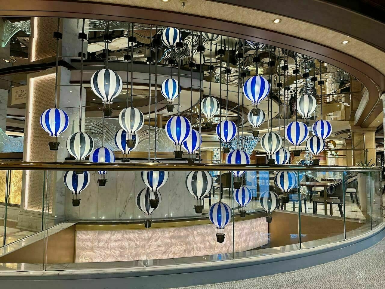 Auto-generated description: The image shows a decorative display of numerous miniature hot air balloons in blue, white, and black, suspended in the interior of a cruise ship